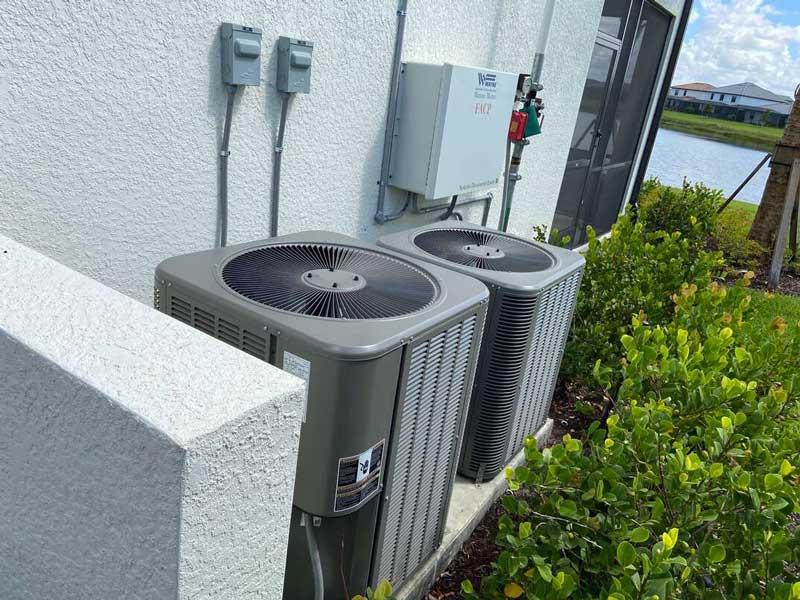 Two outdoor air conditioning units are mounted side by side against the exterior wall of a building. They are surrounded by greenery, including small bushes. The backdrop includes a view of a small water body and some houses in the distance.