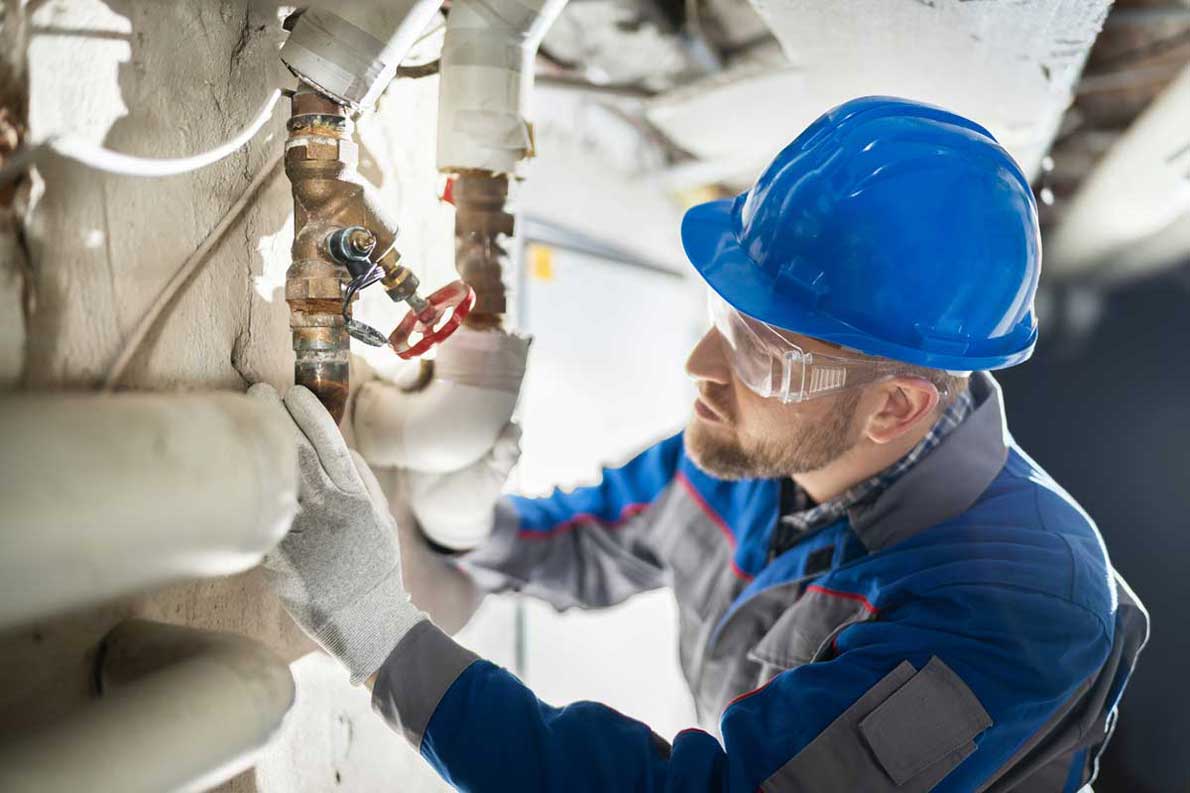 A technician wearing a blue hard hat, safety glasses, and gloves is inspecting and working on pipes in a building. He is focused on a valve and pipes, adjusting and ensuring proper installation. The background shows a typical industrial or utility space.