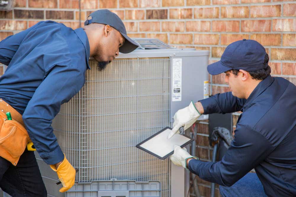 Two technicians wearing navy uniforms and caps work on an outdoor air conditioning unit. One holds a tablet, while the other inspects the unit closely. Both are intent on the task. The brick wall of a building is visible in the background.