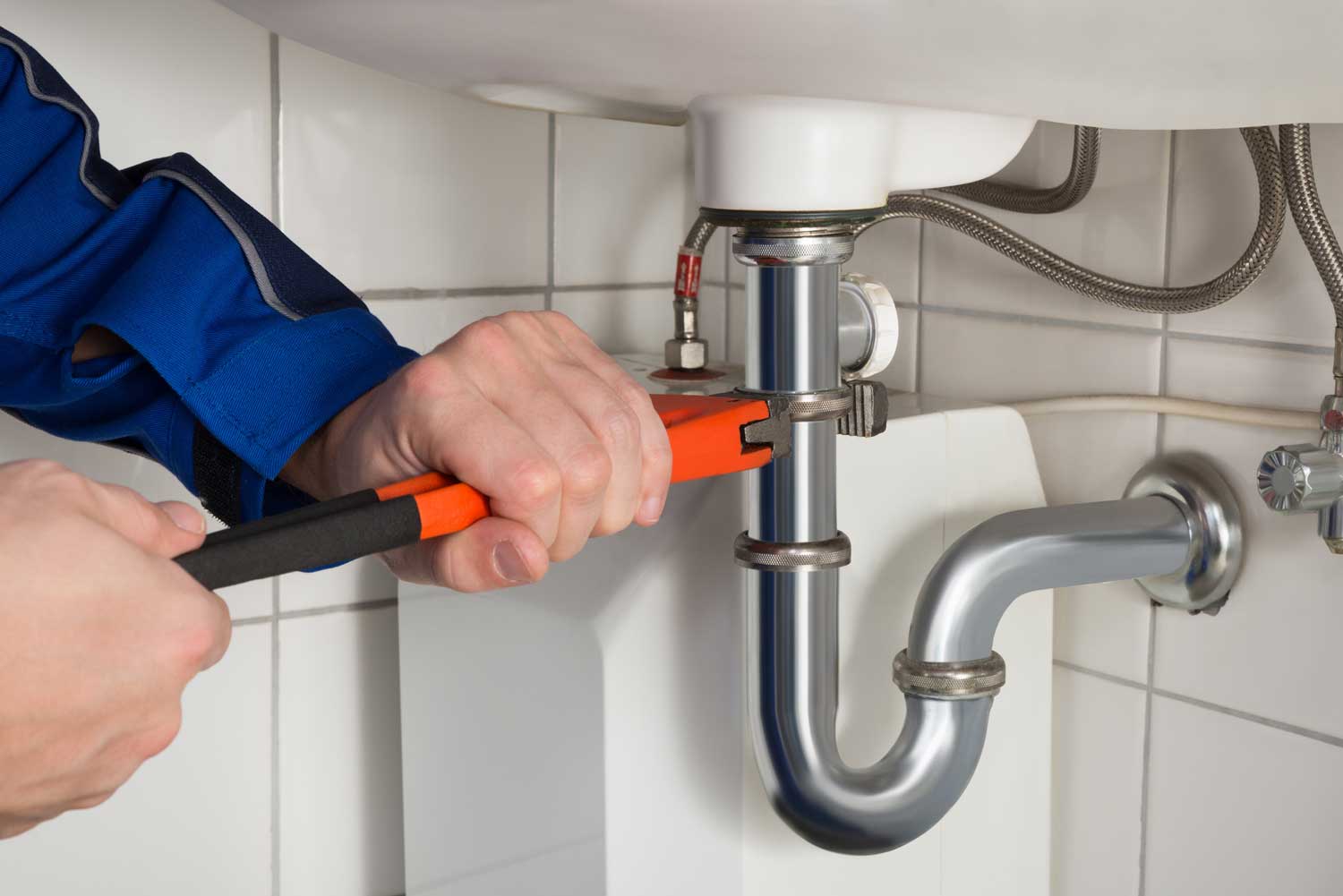 Plumber wearing a blue uniform uses an orange-handled wrench to tighten a pipe under a white ceramic sink in a tiled bathroom. The focus is on the hands working on the plumbing connections, highlighting the tools and metallic pipes.